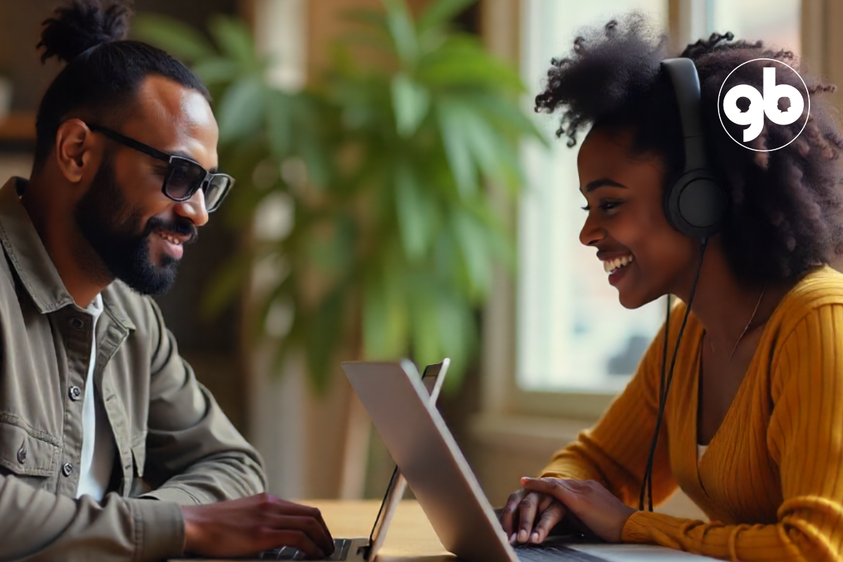 Homem negro com óculos escuros e barba trabalha em frente de uma mulher negra, cada um com seu computador pessoal em uma mesa de trabalho de madeira.