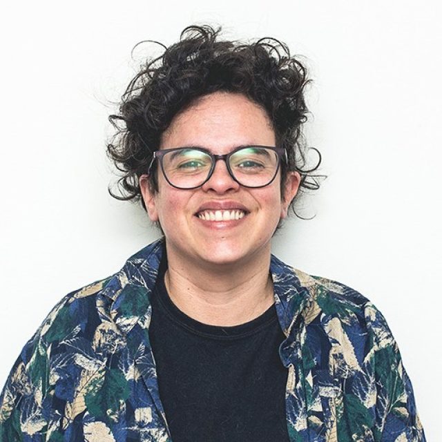 Cintia é mulher branca, de cabelos pretos levemente cacheados. Usa óculos. Veste camisa florida em azul e verde sobre camiseta preta.