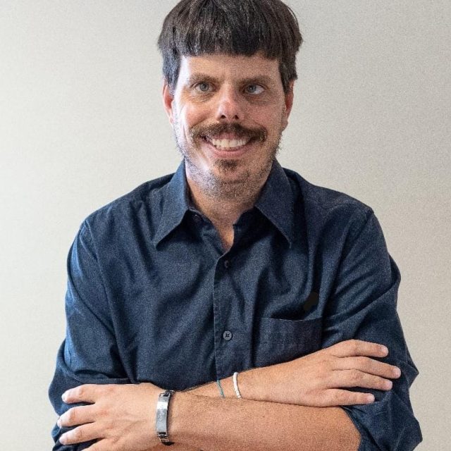 oliver é um homem branco, de cabelos castanhos curtos, olhos claros e com bigode. Veste camisa social azul